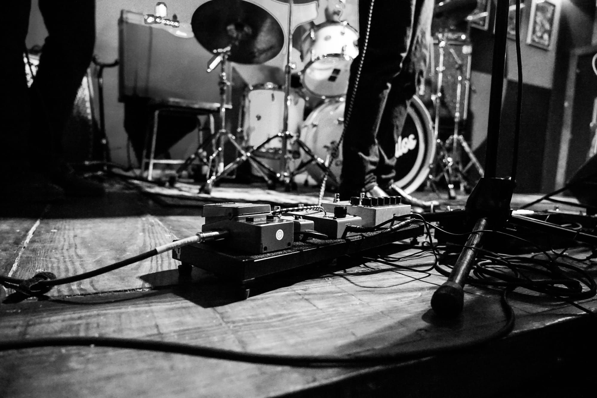 A black and white photo of a small stage: a drum kit, an amplifier and guitar pedals on the floor.