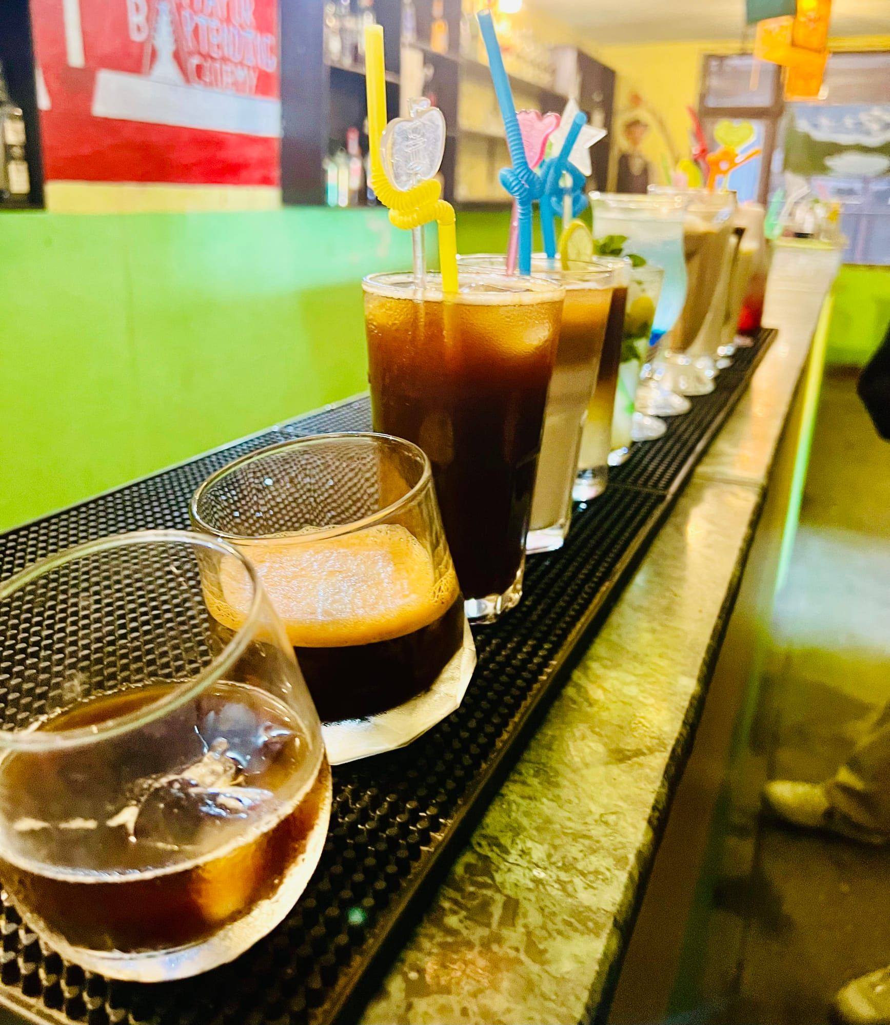 A row of drinks lined up along a bar rail, from dark beers to bright cocktails with straws.