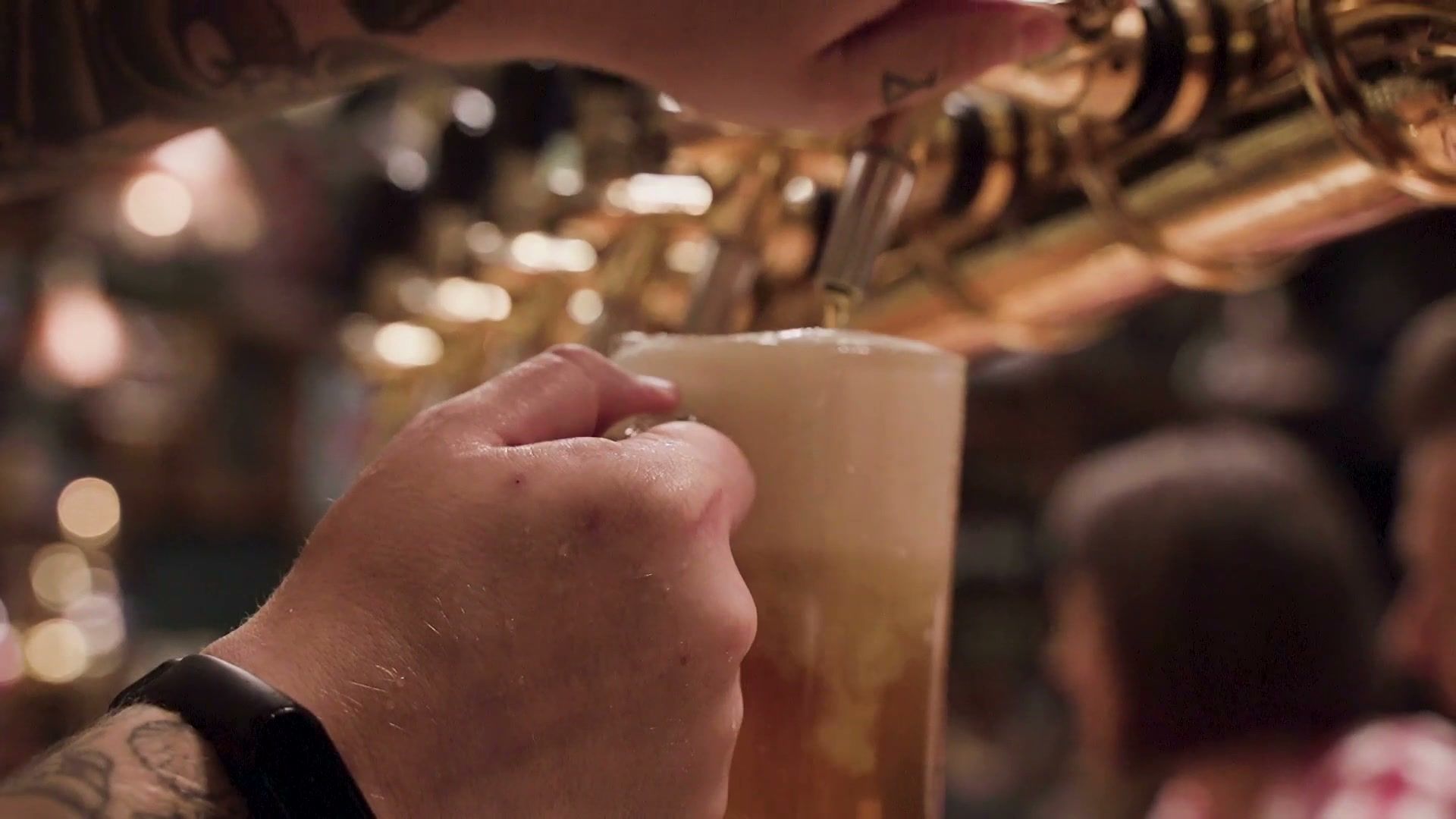 A hand holds a glass mug under a brass beer tap as it fills with foam, with blurred lights and people behind.