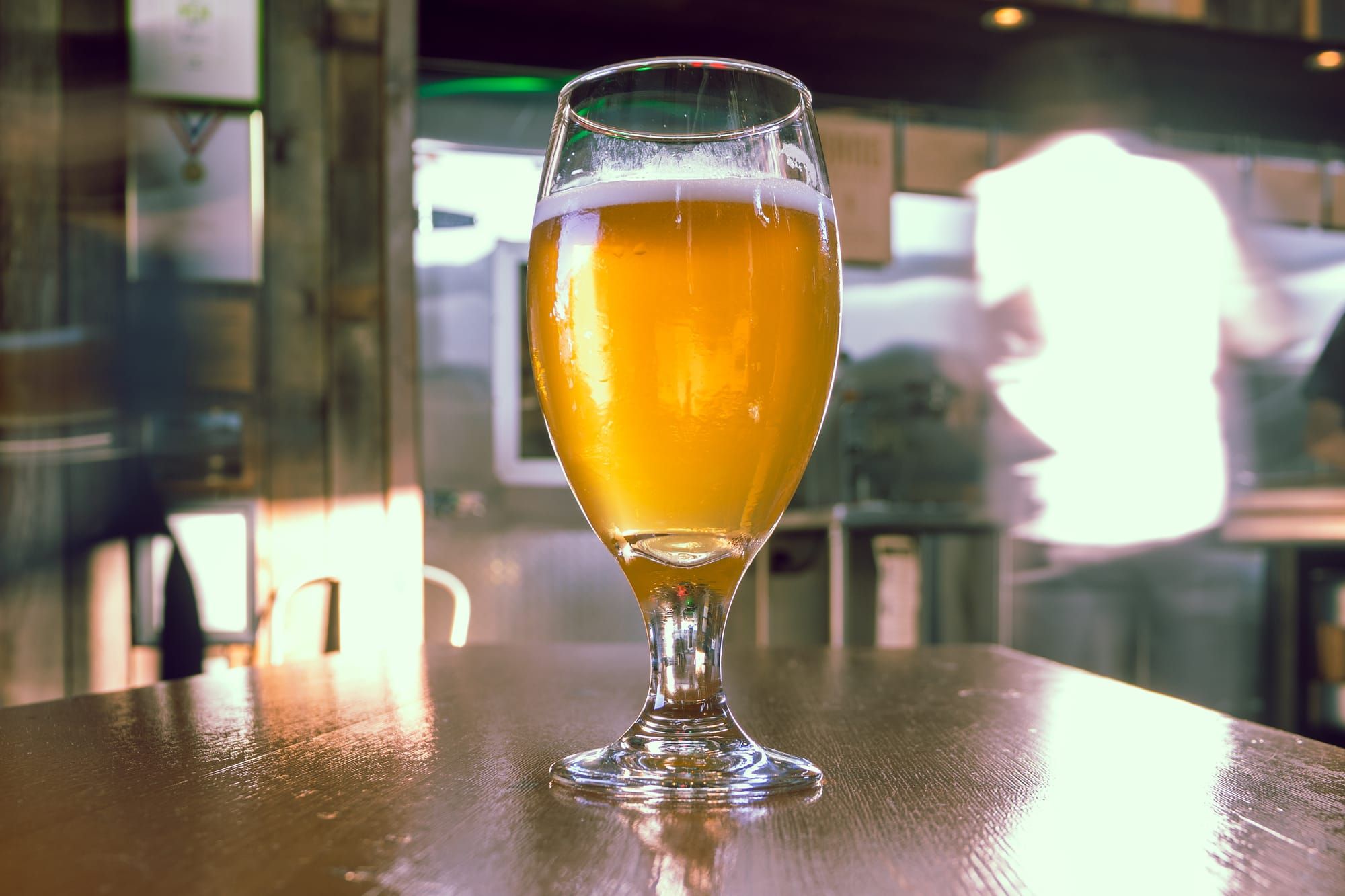 A tulip glass of hazy golden beer on a wooden table in a sunny taproom.