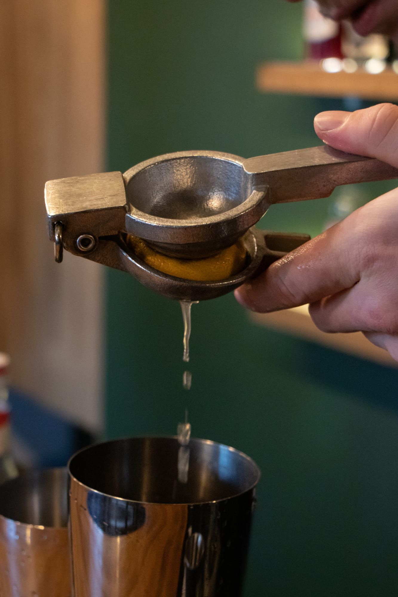 A hand squeezing a lemon through a metal citrus press into a cocktail shaker.